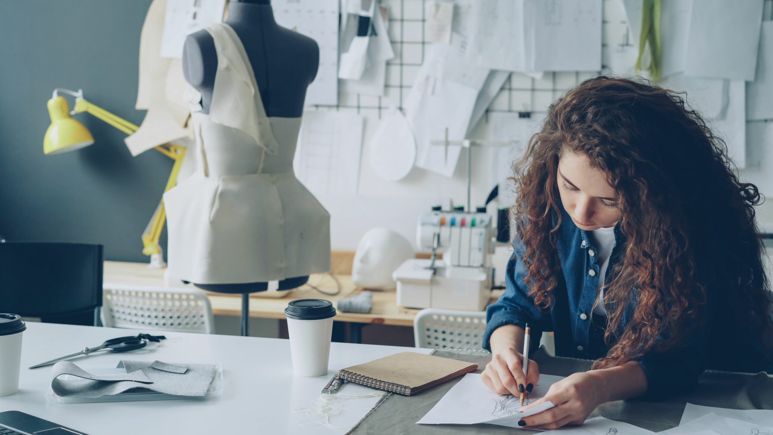 Créatrice de mode dessinant un croquis de vêtement dans son atelier, avec un buste de couturière et des patrons en arrière-plan